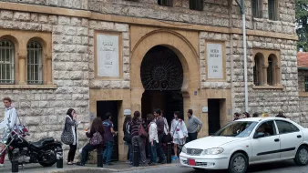 L'image montre l'entrée d'un bâtiment en pierre, probablement une université. Plusieurs personnes sont rassemblées devant l'entrée, créant une petite file d'attente. On peut voir des étudiants, certains portant des sacs. À droite, une voiture blanche passe proche de la scène, tandis qu'une moto est garée à proximité. L'architecture du bâtiment présente des éléments classiques avec un grand portail en fer.