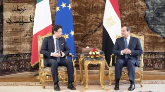 L'immagine mostra due uomini seduti su poltrone dorate in un ambiente elegante. Sullo sfondo si possono notare le bandiere dell'Italia e dell'Egitto, insieme a una bandiera dell'Unione Europea. Entrambi gli uomini stanno conversando e sembrano impegnati in una discussione formale, circondati da elementi decorativi e un tappeto elaborato. Sulle loro ginocchia ci sono bouquet floreali. L'atmosfera è di cordialità e ufficialità.