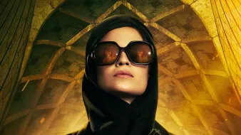 تظهر في الصورة امرأة ترتدي نظارات شمسية وقبعة سوداء، وتحمل مسدسًا. يتميز الخلفية بتصميم معمارية معقدة، حيث تظهر تفاصيل ذهبية وأشكال هندسية، مما يضفي على الصورة طابعًا غامضًا وقويًا. يبدو أن تعبير وجهها يوحي بالثقة والقوة.