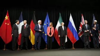 در این تصویر، گروهی از نمایندگان کشورهای مختلف در یک جلسه رسمی ایستاده‌اند. پرچم‌های چین، آلمان، اتحادیه اروپا، ایران، روسیه، بریتانیا و ایالات متحده در پس‌زمینه حضور دارند. هر یک از افراد در لباس‌های رسمی و با حالت‌های متفاوتی در صف ایستاده‌اند، که نشان‌دهنده اهمیت و جدیت جلسه است. به نظر می‌رسد آن‌ها در حال گفت‌وگو و تعامل با یکدیگر هستند.