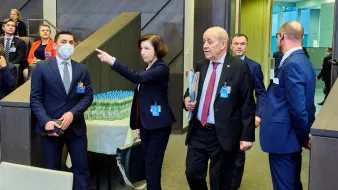 L'image montre un groupe de personnes dans un environnement de conférence ou de réunion. Au centre, une femme portant un blazer indique quelque chose, tandis que d'autres personnes, dont un homme en costume et en cravate, l'écoutent attentivement. Certains participants semblent en discussion, tandis que d'autres regardent dans la direction où la femme pointe. En arrière-plan, on peut voir des bouteilles d'eau sur une table. L'atmosphère semble sérieuse et professionnelle.