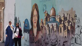 L'immagine mostra due donne che camminano lungo una strada, accanto a un murale. Il murale ritrae una donna con un cappello alla "press" e un volto espressivo. Sullo sfondo, si possono vedere edifici caratteristici, e ci sono scritte in arabo che possono fare riferimento a temi di libertà di stampa o giustizia. La scena è vivace e riflette una cultura ricca e una narrazione importante legata ai diritti dei giornalisti.