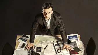 L'image montre un homme debout sur une table en bois, entouré de divers documents et d'objets. Il a une expression sérieuse et semble concentré. Sur la table, on peut voir des cartes, des notes, des dossiers et un lampadaire qui éclaire la scène. L'environnement autour de lui est plutôt sobre, avec des chaises noires et un sol sombre. L'ensemble donne une impression de travail acharné et de recherche.