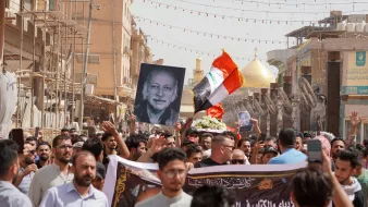 L'immagine mostra una folla di persone durante una manifestazione. Molti manifestanti tengono in alto una grande fotografia di un uomo, insieme a una bandiera irachena. Sullo sfondo si possono vedere elementi architettonici e due cupole dorate, che suggeriscono la presenza di un luogo sacro o importante. L'atmosfera sembra intensa e significativa, con i partecipanti che mostrano il loro sostegno o commemorazione attraverso slogan e simboli.