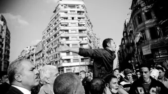 The image depicts a scene of a protest or demonstration. In the foreground, a young man is animatedly speaking or shouting, with his arms outstretched, indicating passion or urgency. Surrounding him are several people, some looking up at him with expressions of engagement or concern. The background features tall buildings, hinting at an urban environment. The photograph is in black and white, which emphasizes the intensity and emotions of the moment. The overall atmosphere conveys a sense of solidarity and activism.