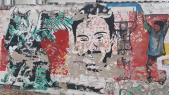 L'immagine mostra un murale su un muro. A sinistra, si può vedere un disegno di una creatura con ali e tratti animaleschi, insieme a elementi astratti e colori vivaci. Al centro, c'è un volto umano stilizzato, con dettagli intricati e un'espressione intensa. Sullo sfondo, si nota un altro personaggio con un gesto di sollevamento delle braccia. La composizione è caratterizzata da una ricca varietà di colori e forme, creando un effetto dinamico e coinvolgente.