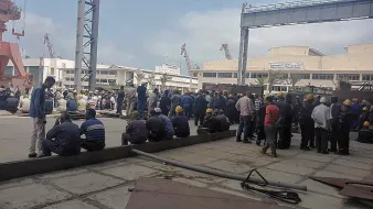 L'image montre un groupe de nombreux travailleurs rassemblés dans un espace extérieur, probablement dans un port ou un chantier naval. Les personnes sont vêtues de tenues de travail et portent des casques de sécurité jaunes. Certains d'entre eux sont assis sur le sol, tandis que d'autres sont debout, discutant ou attendant. L'environnement semble industriel, avec des structures métalliques et des équipements visibles en arrière-plan. Le ciel est partiellement nuageux.