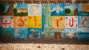L'image montre un mur décoré de peintures colorées. Les motifs incluent des formes géométriques, des fleurs et des éléments abstraits. Les couleurs vives comme le bleu, le vert, le jaune et le rouge créent un contraste attrayant. Le bas du mur semble légèrement abîmé par le temps, et le sol est recouvert de petits débris, ce qui donne un aspect un peu désordonné à l'ensemble. Cette scène évoque un environnement créatif, peut-être dans un espace communautaire ou scolaire.