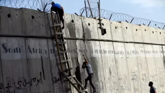 La imagen muestra a dos personas subiendo una pared de concreto alta. Una de ellas está en la parte superior, mientras que la otra está en una escalera de madera, intentando escalar. En la pared hay alambres de púas en la parte superior y también se pueden ver algunas inscripciones y grafitis en varios idiomas. El ambiente parece ser tenso y el contexto sugiere una situación de conflicto o segregación.