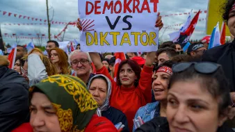L'image montre une foule rassemblée lors d'un événement, probablement une manifestation ou un rassemblement politique. Au premier plan, on voit plusieurs personnes, dont une femme tenant une pancarte sur laquelle est inscrit "DEMOCRAT VS DIKTATÖR". Les participants semblent engagés et déterminés, et les expressions sur leurs visages reflètent des émotions fortes. On peut également observer des drapeaux et des banderoles en arrière-plan, suggérant un contexte de mobilisation politique.
