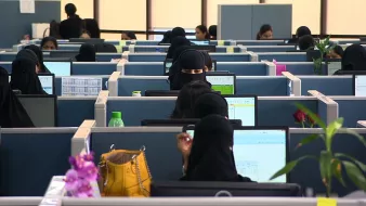 تظهر في الصورة مجموعة من النساء يرتدين ملابس تقليدية، ويعملن في مكاتب داخل بيئة مكتبية. تُلاحظ أن معظم النساء يرتدين النقاب، ويتركز انتباههن على شاشات الكمبيوتر أمامهن. الخلفية تحتوي على مكاتب مقسمة، مع وجود بعض العناصر مثل زجاجات المياه والنباتات. الجو العام يبدو نشطًا ومهنيًا.