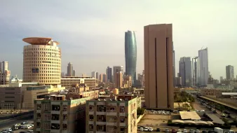 تظهر الصورة منظرًا حضريًا لمدينة حديثة. يمكن رؤية مباني عالية ومتطورة، حيث تبرز ناطحات سحاب زجاجية تعكس الضوء. في المقدمة، توجد مبانٍ ذات تصميمات متنوعة، بعضها قديم Willow وما زال يستخدم. وقد تحتوي المنطقة على مواقف للسيارات ومساحات خضراء بسيطة. السماء تبدو صافية، مما يضفي جوًا هادئًا على المشهد. تعتبر هذه الصورة دليلاً على التمدن والتطور العمراني في المدينة.