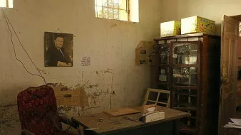 L'image montre une pièce semblant abandonnée ou négligée. Sur le mur, il y a une photo encadrée d'un homme. Le décor comprend une chaise rouge en velours, une table en bois avec quelques objets dessus, et une vitrine en bois remplie de divers éléments. Les murs sont marqués par l'usure et présentent des traces de détérioration. La lumière pénètre à travers une fenêtre, illuminant partiellement l'espace. L'atmosphère générale est nostalgique et témoigne d'un passé révolu.