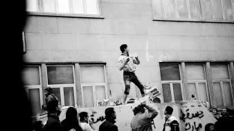 L'immagine mostra una scena di protesta in bianco e nero. Un giovane si trova in cima a un veicolo, apparentemente utilizzando un megafono o uno strumento, mentre sembra incoraggiare la folla sottostante. La gente intorno a lui è visibilmente coinvolta, alcuni alzano le mani o applaudono. Sul veicolo ci sono delle scritte, e l'architettura sullo sfondo suggerisce un contesto urbano. L'atmosfera è carica di energia e di determinazione.