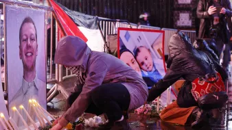 تصویر فضای یک تجمع یادبود را نشان می‌دهد که در آن افراد در حال قرار دادن شمع و گل هستند. در جلوی آنها، تصاویری از یک فرد و یک خانواده به نمایش گذاشته شده است. باران به زمین می‌ریزد و خیابان براق است. افراد با لباس‌های گرم و کلاه روی زمین نشسته‌اند و به یاد افرادی که از دست رفته‌اند، مراسمی برگزار می‌کنند. این صحنه حال و هوای غم و اندوه را منتقل می‌کند.