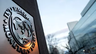 L'image montre le logo du Fonds monétaire international (FMI) en premier plan, avec un globe stylisé et des feuilles. En arrière-plan, on aperçoit un bâtiment moderne, probablement le siège du FMI, baigné dans une lumière douce du crépuscule. Le ciel est clair, avec des nuages légers. L'ensemble évoque une ambiance institutionnelle et internationale.