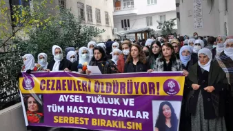 L'image montre un groupe de personnes rassemblées dans une rue, tenant des pancartes et des banderoles. Les femmes portent des foulards sur la tête et semblent soutenir une cause ou un mouvement. L'une des banderoles porte des slogans demandant la libération d'Aysel Tuğluk et des prisonniers malades. Le contexte semble être une manifestation pour les droits de l'homme et la justice. Des expressions de détermination et de solidarité sont visibles parmi les participants.