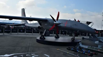 The image features a military drone displayed prominently on a platform. The drone has a sleek, aerodynamic design with multiple red propellers. It is painted in a gray color, which is common for military equipment. In the foreground, there are several missiles and munitions arranged neatly, showcasing the drone's armament potential. The background includes a tent structure and additional drone models, suggesting this is part of an exhibition or airshow. The branding on the drone indicates it is a product of Baykar, referencing a specific model or variant likely designated for military use.