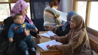 L'image montre une scène dans un environnement communautaire. On peut voir une femme portant un foulard qui tient un bébé dans ses bras, s'approchant d'une table où une autre femme, également couverte d'un foulard, est assise et prend des notes. À l'arrière-plan, d'autres femmes semblent occupées à diverses activités. L'ambiance semble être celle d'un événement organisé, possiblement une inscription ou une réunion. Les couleurs et les vêtements des femmes reflètent une culture riche et diversifiée.