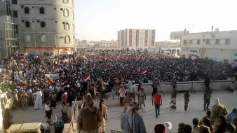 الصورة تظهر تجمعًا حاشدًا من الناس في مكان عام، حيث يبدو أنهم يشاركون في فعالية أو تظاهرة. المجموعة كبيرة وتحتوي على عدد من الأعلام، مما يشير إلى أن هناك موضوعًا معينًا يجمعهم. المباني المحيطة توحي بأن المكان في منطقة حضرية، كما أن بعض الأشخاص يجلسون على الدرج بينما يتواجد آخرون في المقدمة. تبرز الأجواء الحماسية في الحشد.