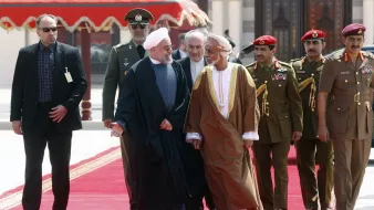 The image features a group of men walking together on a red carpet, likely in a formal or diplomatic setting. In the foreground, two men are engaged in conversation, one wearing a black suit and the other in traditional attire. They appear to be leaders or dignitaries. Behind them, there are several men in military uniforms and suits, possibly security personnel or officials, accompanying the two main figures. The background suggests an ornate architectural setting, enhancing the official nature of the gathering.