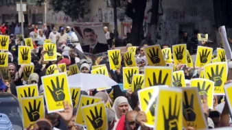 L'image montre une grande foule de personnes participant à une manifestation. Elles tiennent des affiches jaunes avec un symbole de main noire en faisant le signe qui pourrait représenter un mouvement ou une cause spécifique. La foule semble déterminée et engagée, et l'atmosphère est chargée d'énergie. On peut voir des pancartes et des slogans en arrière-plan. L'événement semble vouloir exprimer une opinion ou revendiquer des droits.