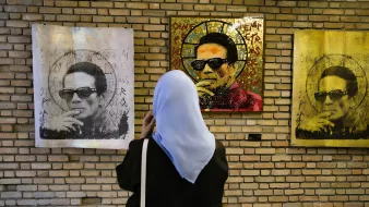 Nell'immagine si vede una persona con un velo che osserva attentamente delle opere d'arte appese a una parete di mattoni. Le opere raffigurano un uomo con occhiali da sole e un'espressione seria. I quadri presentano colori vivaci e schemi di texture, creando un contrasto interessante con lo sfondo neutro dei mattoni. L'ambiente sembra essere una galleria d'arte, con un'atmosfera contemplativa.