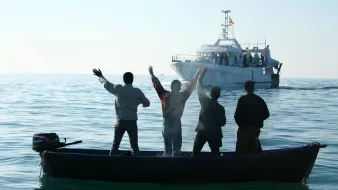 The image depicts four individuals in a small boat on calm waters, facing away from the camera. They are raising their arms, seemingly in a gesture of celebration or greeting. In the background, a larger boat is approaching, with several people visible on it. The scene is set against a clear sky, suggesting a peaceful day on the water.