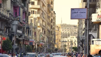 L'image montre une rue animée remplie de voitures et de passants. On peut voir des bâtiments de plusieurs étages de part et d'autre de la rue, avec des balcons et des façades variées. La scène semble se dérouler pendant la journée, la lumière du soleil illuminant l'environnement urbain. Des panneaux de signalisation et des enseignes en arabe sont visibles, ajoutant à l'atmosphère locale de cette ville. Les gens semblent se déplacer rapidement, créant une ambiance dynamique et vivante.