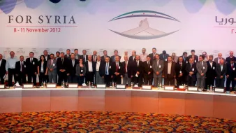 L'image montre un groupe de personnes réunies lors d'un événement formel, probablement une conférence. Elles sont alignées en plusieurs rangées, toutes en costume, et semblent participer à un rassemblement lié à la Syrie, comme indiqué par le texte en arrière-plan. Le décor est coloré, avec des drapeaux et des motifs qui évoquent l'événement. Les participants adoptent des expressions sérieuses, ce qui suggère l'importance de l'occasion.