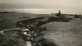 L'image représente un champ de bataille avec une tranchée. On peut voir un soldat assis sur les bords de la tranchée, qui semble surveiller les environs. Le terrain est accidenté et semé de sacs de sable, typiques des fortifications de la Première Guerre mondiale. À l'arrière-plan, on aperçoit une étendue d'eau qui pourrait être un lac ou une rivière. L'atmosphère générale est sombre et mélancolique, évoquant la dureté de la guerre.