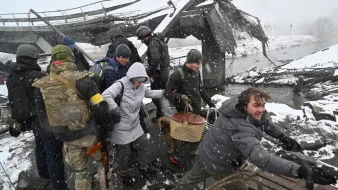 L'immagine mostra un gruppo di persone in un'area distrutta, probabilmente a causa di un conflitto o di un disastro. Alcune persone indossano abbigliamento militare, mentre altre sembrano civili. È in corso un salvataggio o un'evacuazione, con qualcuno che sta aiutando un'altra persona a attraversare il terreno difficile. La scena è caratterizzata da neve e condizioni atmosferiche difficili, suggerendo un ambiente critico e di emergenza. I volti mostrano preoccupazione e determinazione.