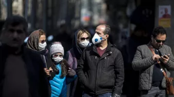 The image depicts a group of people walking on a city street while wearing face masks. Among them, a man is wearing a blue mask, and there is a girl in a blue vest. Everyone appears to be maintaining a level of social distance. In the background, the scene is somewhat blurred, implying activity and movement in an urban environment. Some individuals are engaged in their own activities, such as checking their phones. The overall mood suggests a public setting where health precautions are being observed.
