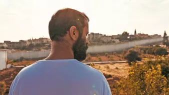 L'image montre un homme vu de dos, contemplant un paysage. Il a une barbe et porte un t-shirt blanc. En arrière-plan, on aperçoit des bâtiments sur une colline, flanqués d'une barrière ou d'un mur. La lumière est douce, créant une atmosphère paisible, et le ciel présente quelques nuages. Le terrain est principalement sec avec des touches de verdure autour.
