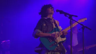 L'immagine mostra un musicista sul palco, intento a suonare una chitarra elettrica di colore azzurro. Ha i capelli ricci e indossa una camicia ampia con un look casual. Dietro di lui si intravedono altri musicisti e luci bluastre che creano un'atmosfera suggestiva. L'artista sembrano concentrato e coinvolto nella sua performance.