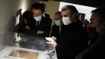 L'image montre un groupe de personnes, dont une figure centrale qui semble être un homme en train d'observer des documents exposés dans une vitrine. Ils portent tous des masques, probablement en raison de mesures sanitaires. L'environnement est sombre, avec des murs clairs, et il y a un intérêt évident pour les objets présentés. Les autres personnes dans le cadre semblent également attentives et engagées dans la discussion.