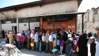 در تصویر، گروهی از مردم به صف ایستاده‌اند. ساختمان در پس‌زمینه دارای نمای ساده‌ای است و تابلو بزرگی بالای درب ورودی نصب شده است. افراد در صف شامل مردان و زنانی هستند که برخی از آن‌ها سبدها یا کیسه‌های خرید را در دست دارند. نور خورشید قوی به تصویر تابیده و فضا شلوغ به نظر می‌رسد. برخی از افراد به طرف دوربین نگاه می‌کنند و برخی دیگر مشغول صحبت هستند. دوچرخه‌ها نیز در کنار خیابان به چشم می‌خورند.