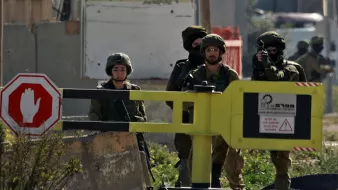 L'immagine mostra un gruppo di soldati in un ambiente urbano. Sono vestiti con uniformi militari e indossano elmetti. Due soldati sono in piedi vicino a un blocco stradale giallo con un segnale di fermo e il gesto di "stop". Nel gruppo, uno dei soldati sta puntando un'arma in direzione non visibile. Il contesto sembra suggerire un'operazione di sicurezza o un controllo di accesso. Sullo sfondo, ci sono strutture edilizie e segni di una zona di conflitto.