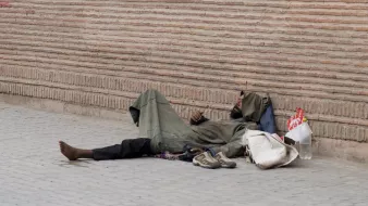الصورة تظهر شخصًا ممددًا على الأرض، يرتدي ثيابًا بسيطة وفضفاضة. يبدو أنه في حالة استرخاء أو نوم. بجانبه بعض الأغراض مثل حذاء وحقيبة. الخلفية تتكون من جدار مصنوع من الطوب، مما يضيف إلى الأجواء الحضرية للصورة. المشهد يعكس حالة التحدي والمعاناة في الحياة اليومية لبعض الأفراد.