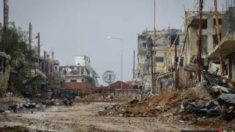 L'image montre une scène de destruction urbaine. On peut voir des bâtiments en ruines, avec des murs effondrés et des structures endommagées. Le sol est recouvert de débris, et l'atmosphère semble sombre et désolée. Il y a des poteaux électriques dans l'arrière-plan, et un éclairage public est visible au loin, mais tout cela contribue à une ambiance de désolation. La végétation est rare, et l'ensemble dégage une impression de lieu abandonné et ravagé.