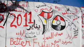 L'image montre un mur recouvert de graffitis en arabe et en anglais. Parmi les inscriptions, on trouve des références à l'Égypte, à la révolution de 2011, et des slogans tels que "Meilleur avenir" et des phrases évoquant la détermination et l'espoir. Les couleurs rouge, noir et jaune du drapeau égyptien sont visibles, renforçant le message patriotique et engagé. L'ensemble évoque une période de changement et de revendications sociales.