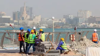 L'image montre un groupe de travailleurs sur un chantier de construction près de la mer. Ils portent des vêtements de sécurité, comme des gilets réfléchissants et des casques. En arrière-plan, on peut voir une ville avec des bâtiments modernes et des structures élevées. L'ambiance semble occupée, et le chantier est environné de matériaux de construction, avec des blocs empilés. La mer est visible au loin, ajoutant un cadre pittoresque à la scène.