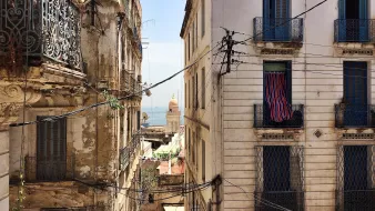 L'image montre une vue d'une rue étroite et animée d'une ville méditerranéenne. De chaque côté, on voit des bâtiments anciens avec des façades usées et des balcons en fer forgé. Des fils électriques serpentent entre les immeubles. Au loin, on peut apercevoir un dôme qui pourrait appartenir à une église ou une mosquée, et un bout de mer est visible, ajoutant une touche de bleu au paysage. Les fenêtres des bâtiments sont ornées de volets ou de rideaux, et l'ensemble dégage une atmosphère nostalgique et pittoresque.