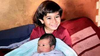 Nell'immagine si vede un bambino più grande che sorride mentre tiene in braccio un neonato avvolto in una coperta blu. Il bambino più grande ha i capelli scuri e lunghi, e indossa una maglietta rossa. Il neonato ha i capelli scuri e un'espressione curiosa. Sullo sfondo ci sono delle coperte sul lettino, creando un'atmosfera accogliente e familiare.