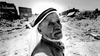L'image montre un homme âgé au visage marqué, qui regarde directement vers l'objectif. Il porte un keffieh, un couvre-chef traditionnel, et a une expression qui dégage une certaine sagesse ou tristesse. En arrière-plan, on peut apercevoir des ruines, des débris et des bâtiments partiellement détruits, suggérant un environnement difficile ou une situation de conflit. La scène est capturée en noir et blanc, ce qui accentue les textures et les contrastes des éléments environnants.