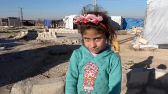 La imagen muestra a una niña pequeña en un campo de refugiados. Ella tiene largas pestañas y ojos claros, y lleva una sudadera de color azul con un diseño en la parte delantera. En su cabeza, tiene una diadema decorada con flores. El entorno es árido, con estructuras de refugio improvisadas y un cielo despejado. La expresión de la niña es curiosa y ligeramente sonriente.