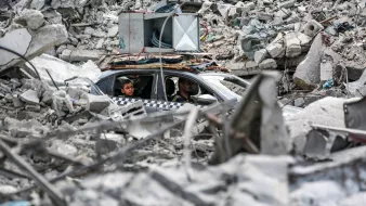 L'immagine mostra una scena di distruzione, con un'ampia quantità di macerie sparse ovunque. Al centro, c'è un'auto parcheggiata tra i detriti, con due persone sedute all'interno. Le macerie sembrano provenire da edifici crollati, evidenziando un contesto di crisi o di conflitto. L'atmosfera è carica di tensione e desolazione, con un forte contrasto tra l'umano e la devastazione circostante.