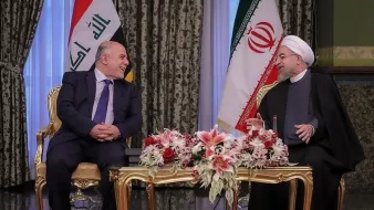 تظهر الصورة لقاءً بين شخصين يجلسان على طاولة مزينة بزهور. العلم العراقي وعلم إيران خلفهم، مما يشير إلى أن الاجتماع يعقد بين ممثلين عن البلدين. يبدو أن كلا الشخصين يستمتعان بالنقاش ولديهما تعابير وجه إيجابية. الديكور في الخلفية أنيق ومزخرف، مما يعكس طابعًا رسميًا لهذا اللقاء.