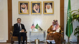 Nell'immagine si possono vedere due uomini seduti uno di fronte all'altro in un ambiente formale. Sullo sfondo ci sono tre ritratti incorniciati, probabilmente di leader sauditi. A sinistra, è presente una bandiera americana e a destra una bandiera saudita. Il tavolo tra i due uomini contiene alcuni oggetti decorativi. L'atmosfera apparente è di dialogo politico o diplomatico.