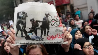 L'immagine mostra una manifestazione con un gruppo di persone unite in segno di protesta. In primo piano, una persona tiene in alto un giornale che riporta un'immagine significativa legata all'evento. La folla sembra esprimere emozioni forti, con alcune donne visibilmente agitatesi e altre che mostrano determinazione. Gli striscioni e le espressioni nei loro volti indicano il loro impegno per una causa importante. Il contesto generale evoca tensione e partecipazione attiva a una questione sociale.